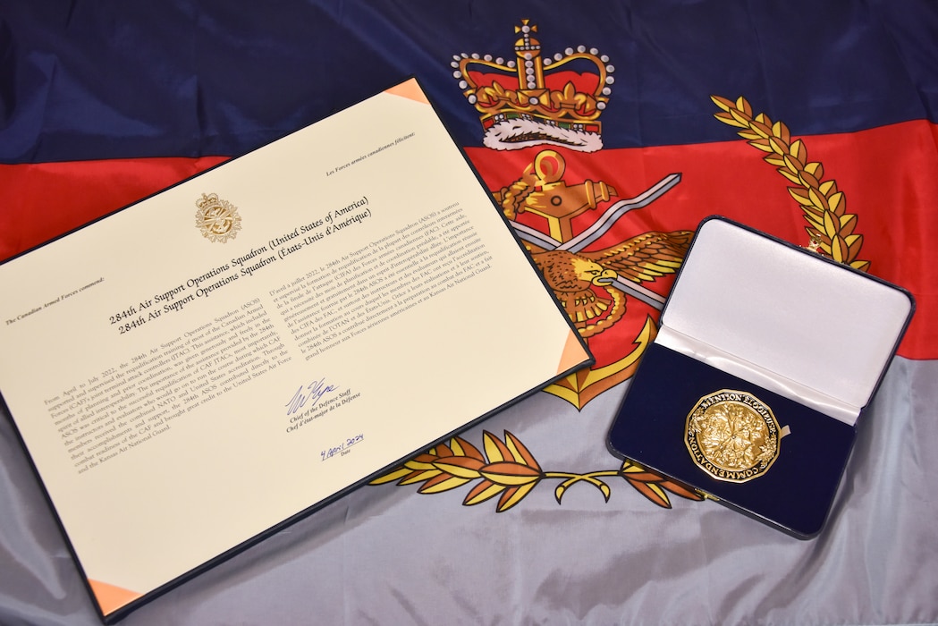 A twelve-sided gold-colored medallion bearing the symbols of the three services: the anchor, the crossed swords, and the flying eagle topped by the royal crown, is displayed in a box next to an award citation on top of a special commemorative flag.