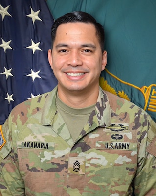 Man in U.S. Army uniform standing in front of two flags.