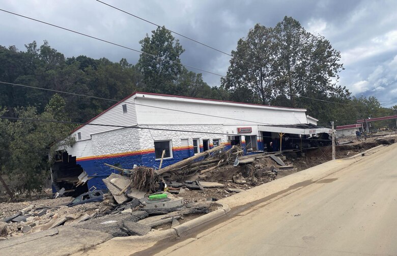 A damaged flood damaged building sits along side a roadway. Debris is piled against the building