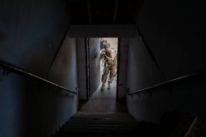 A dark stairwell pointed toward an open door with a military man blasting through.