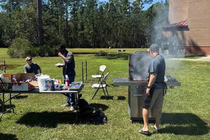 A person grills outdoors while two people chat near a full picnic table.