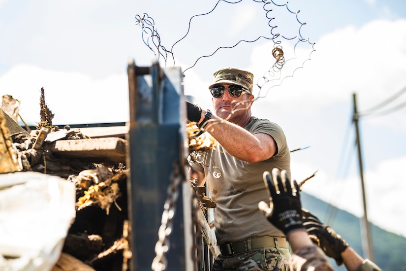 An airman clears debris from atop a metal wall during the day.