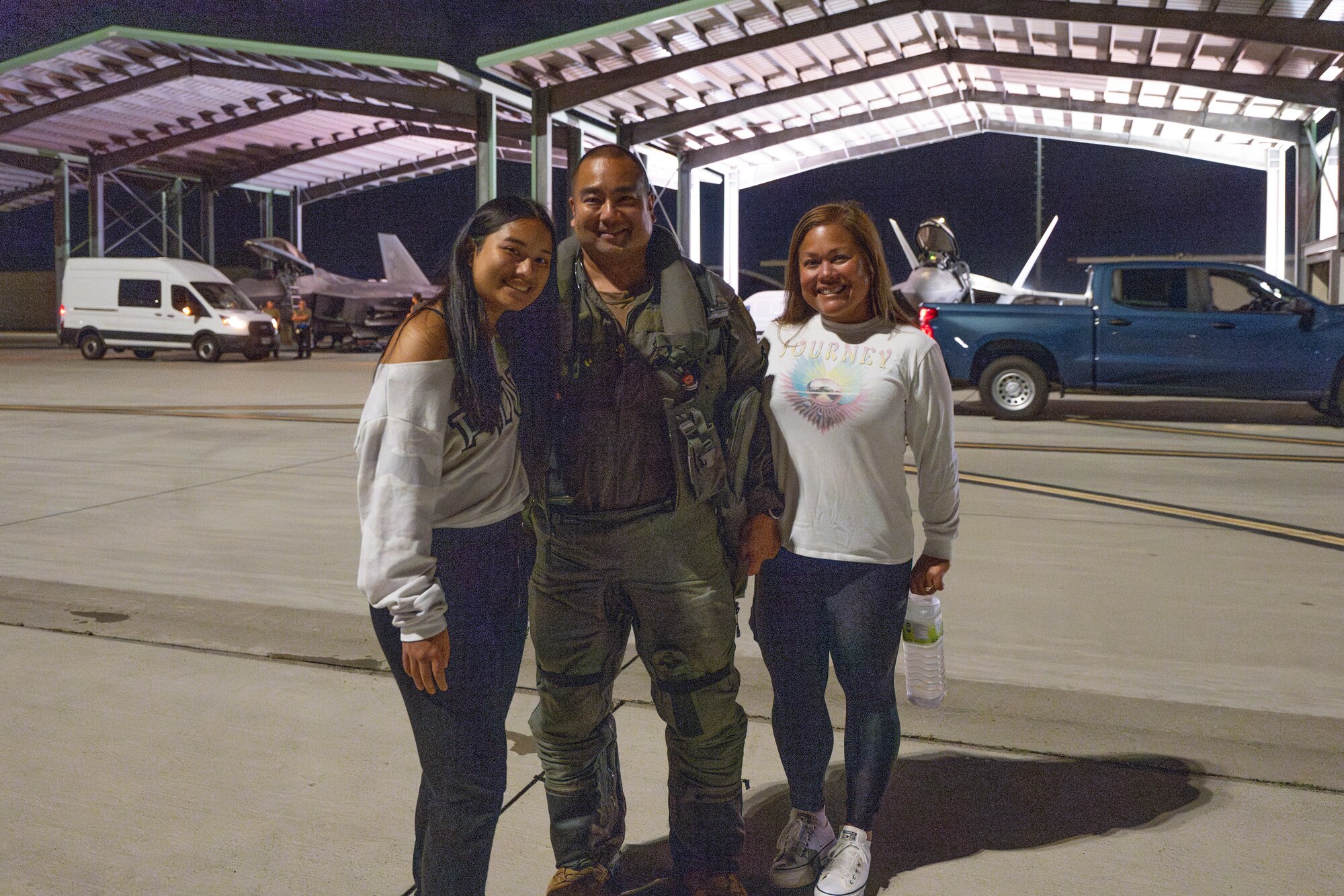A pilot poses with his family at night in front of a hangar.