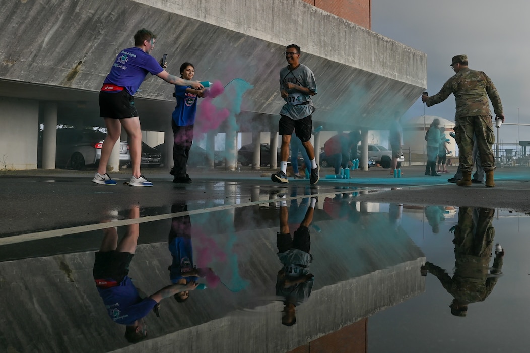 CDC suicide prevention - Photo of the action happening but also a reflection in a nearby puddle of an Airman running through a blast of colored powder being thrown by participants near the finish line of the color run race.