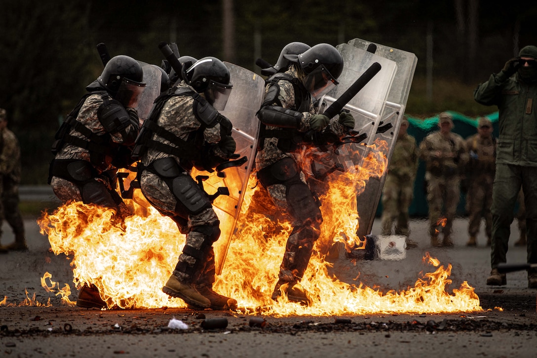 Soldiers wearing riot gear and carrying nightsticks and shields step into flames burning around them.