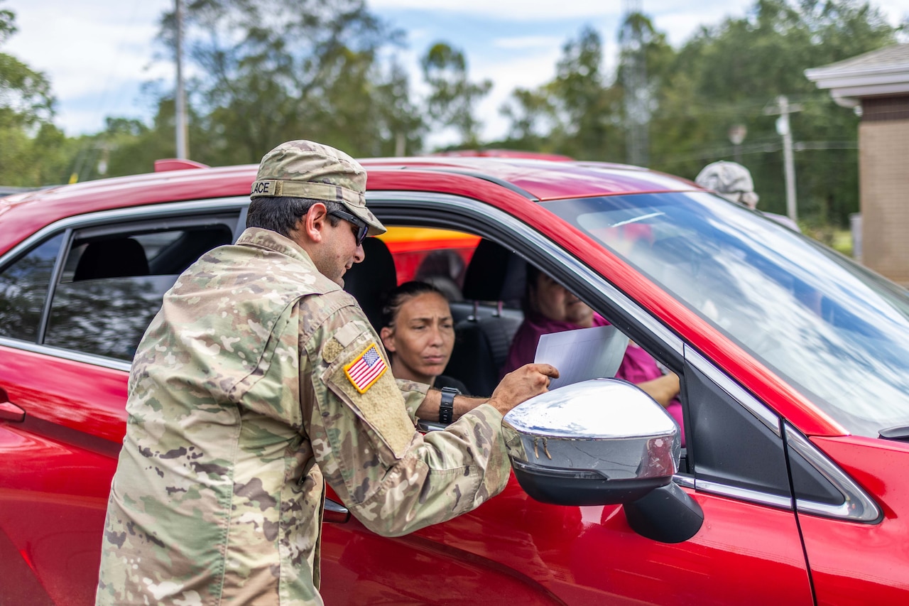 A soldier stands at the passenger window of a car holding a sheet of paper while talking to the people inside the car.