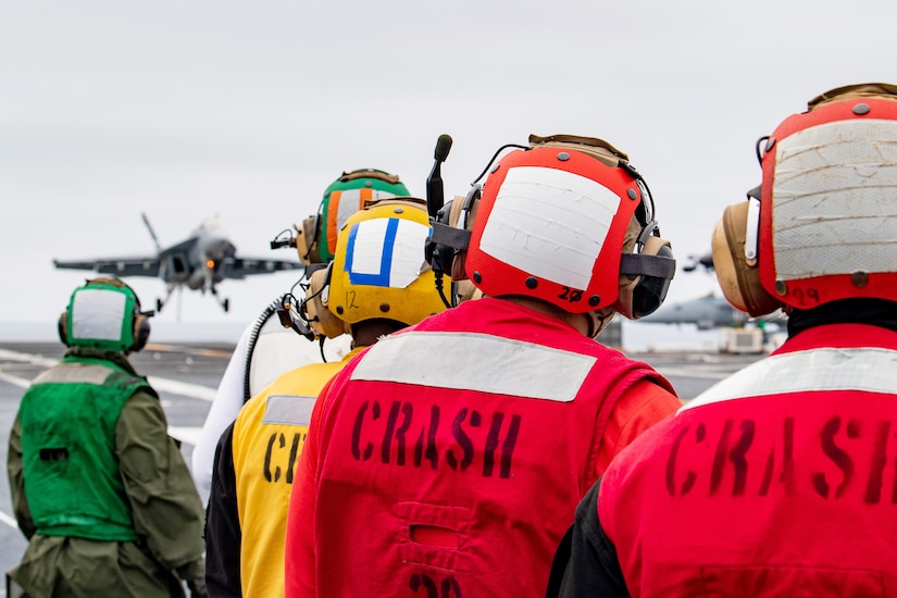 Five sailors wearing colorful clothing watch as an aircraft lands aboard a ship at sea on a cloudy day.