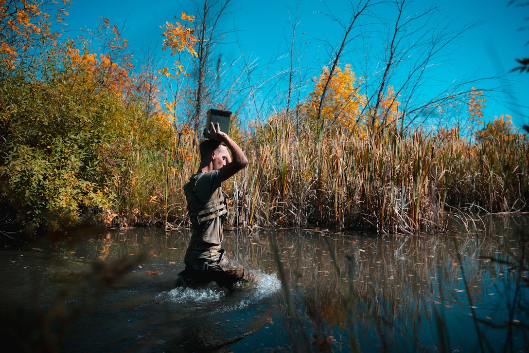 A service member carries an ammunition box on their head while kneeling in a creek surrounded by tall grass under a blue sky.