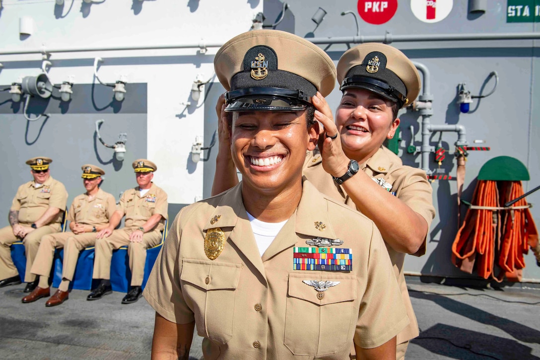 A sailor places a hat on a fellow smiling sailor's head as three seated sailors watch from the left aboard a ship.