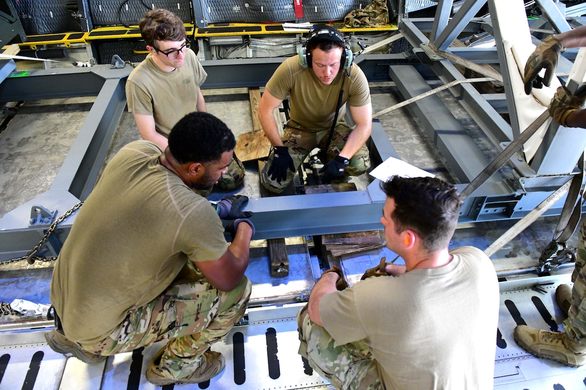 Men in military uniform strapping down metal parts inside aircraft.