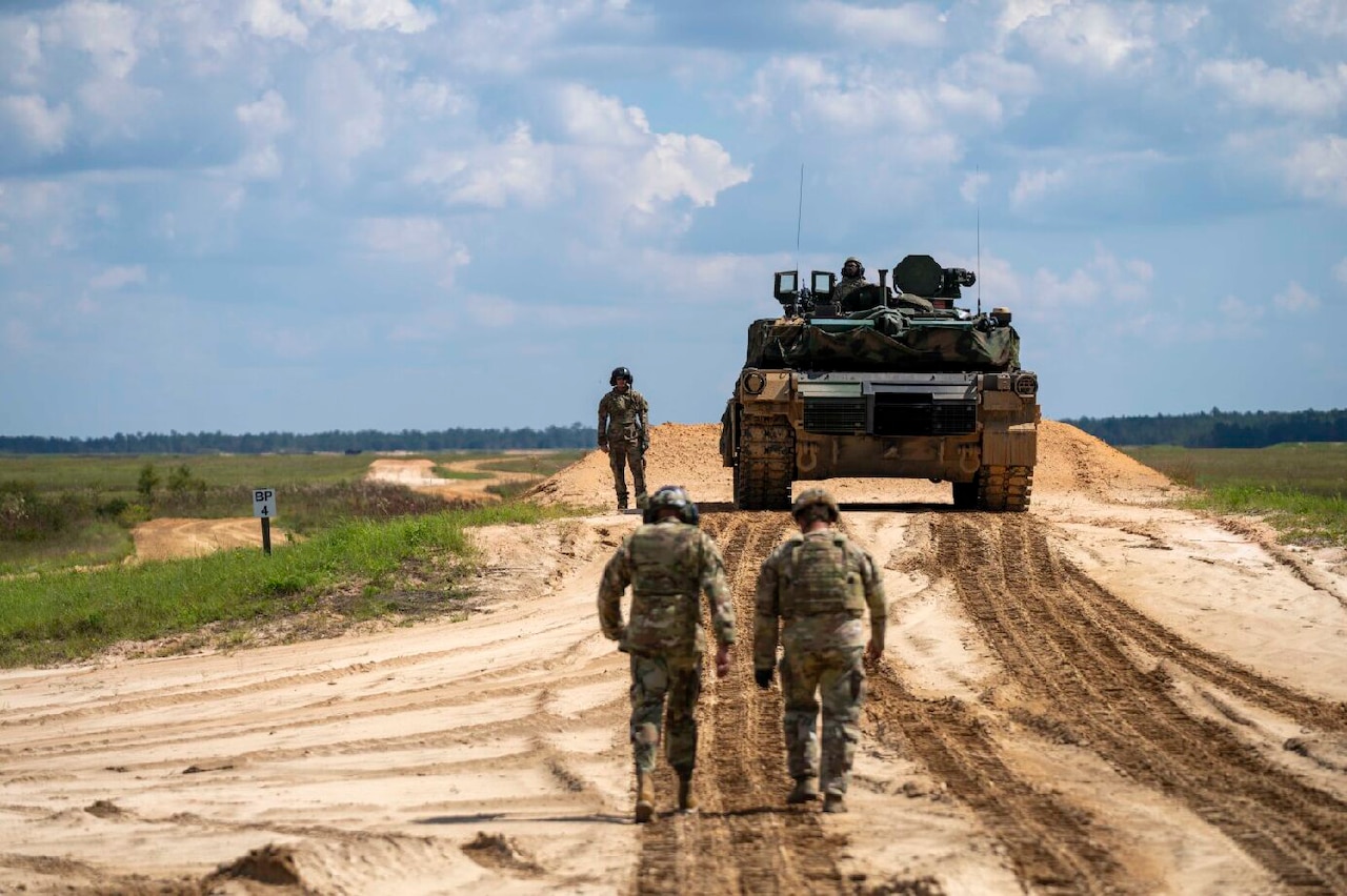 Military personnel walk on a dirt road in the direction of a military tank.