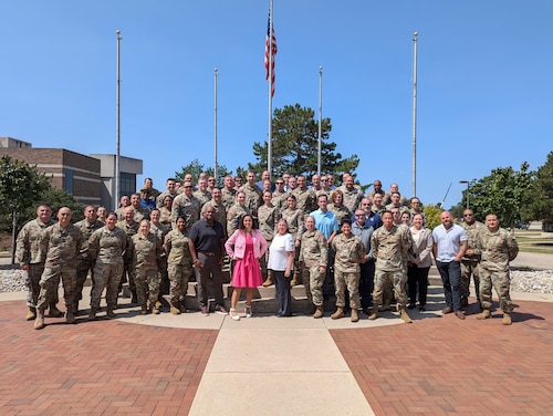 A group of military and civilians standing in front of an American Flag