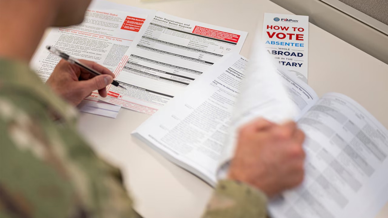 A service member turns the page of a book on a desk while holding a pen.