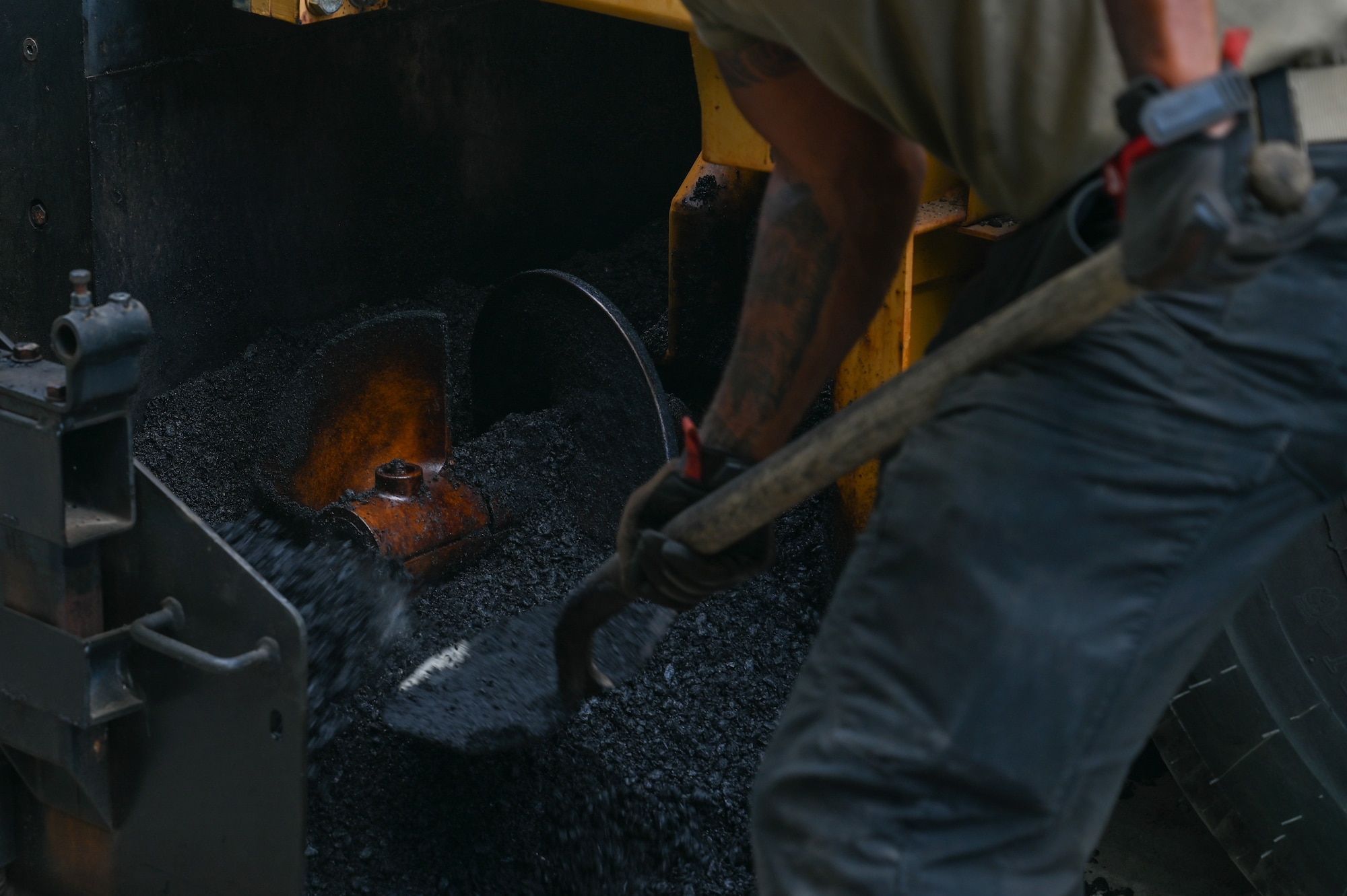 medium shot photo of an airmen shoveling asphalt in part of a machine that lays down asphalt.