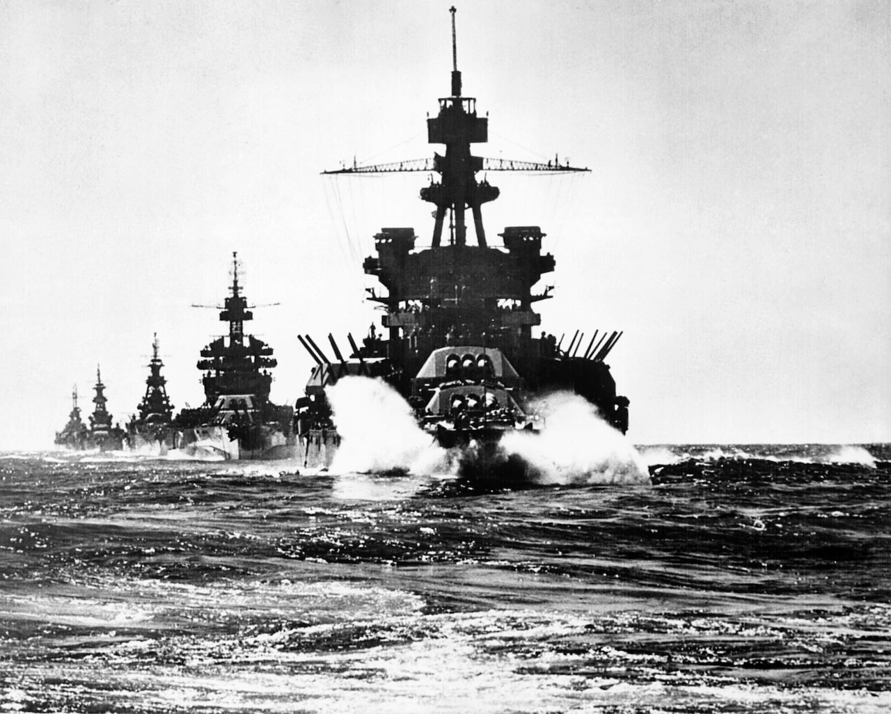 A battleship leads a line of other Navy ships at sea.