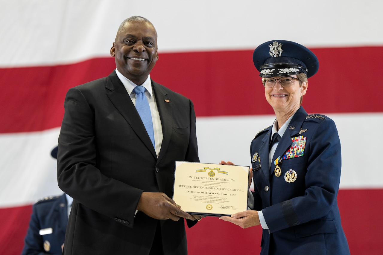 Two people smile while holding an award in front of an American flag.