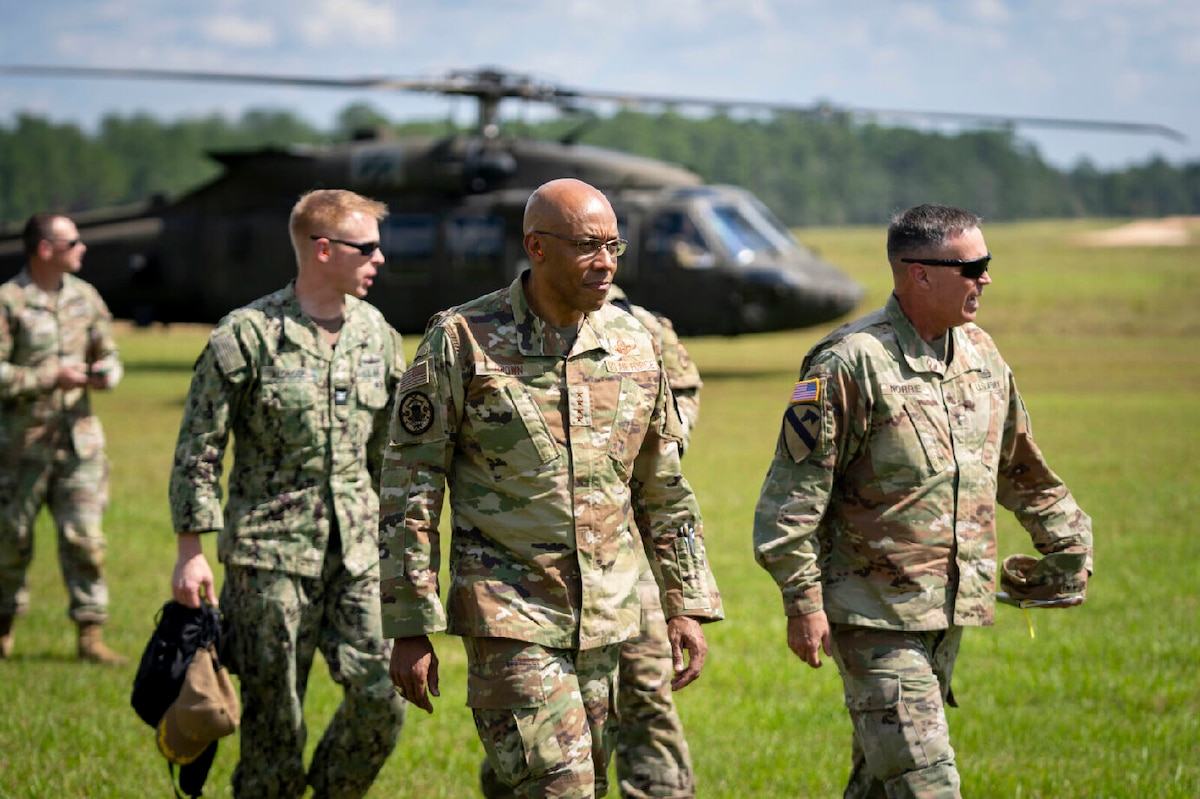Five military personnel walk in a field during daylight. A military helicopter is in the background.