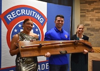 3 Army Soldiers in civilian attire smile while holding a large award in front of a Recruiting Brigade backdrop