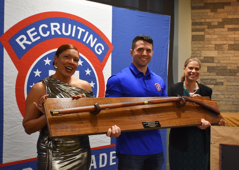 3 Army Soldiers in civilian attire smile while holding a large award in front of a Recruiting Brigade backdrop