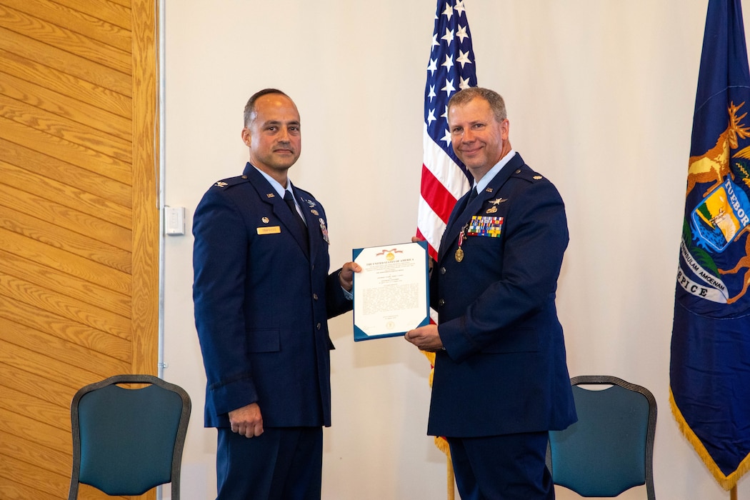 Two male Air Force officer in dress blue uniforms, hold award citation between them looking at camera.