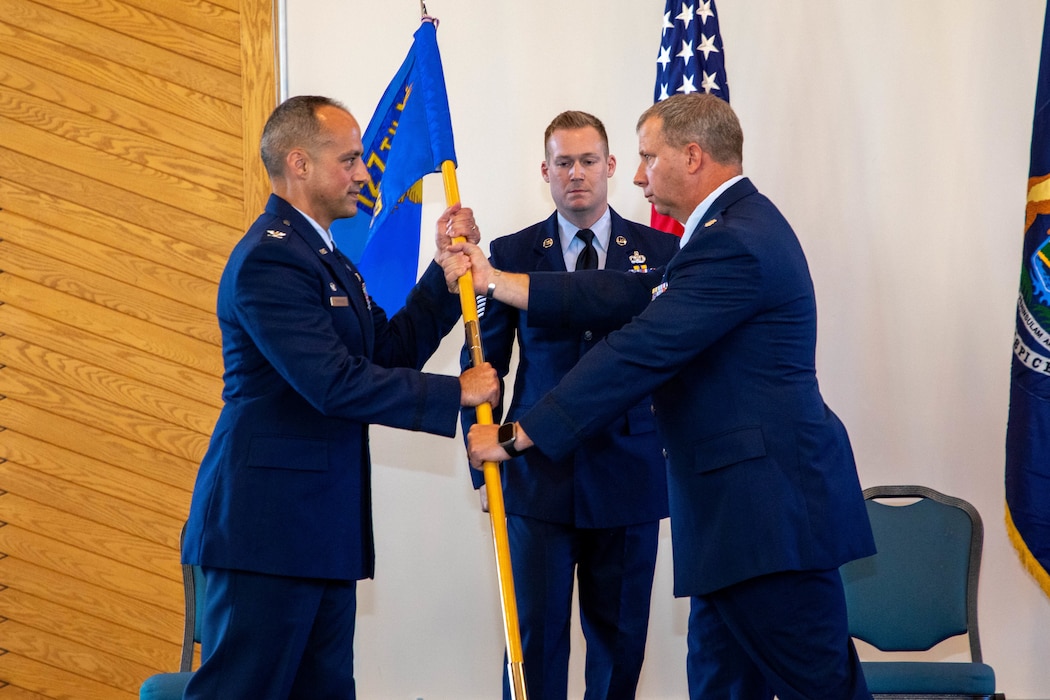 Male Air Force officer in dress blue uniform, hands unit flag to male Air Force officer in dress blue uniform while male non commissioned officer in dress blue uniform stands behind.