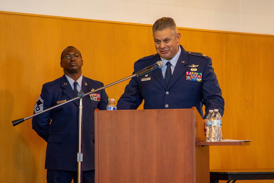 Male Air Force officer in dress blue uniform, stands behind podium and speaks while male Air Force non-commissioned officer stand behind in dress blue uniform.