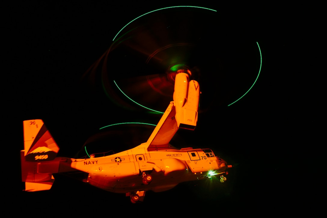 A military aircraft bathed in orange light takes off from a ship at night. The aircraft’s propeller lights create two green rings in the darkness.