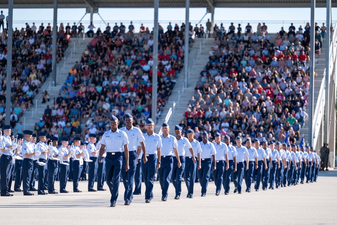 Airmen walk in formation in a stadium with crowds of people in the stands. Other airmen stand in formation to one side holding musical instruments.