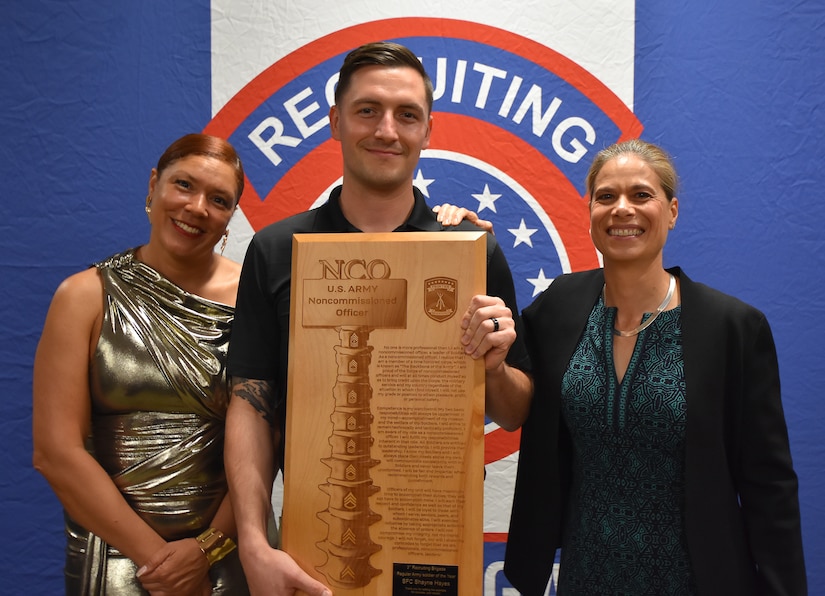 Three Soldiers pose with an award plaque in front of an Army Recruiting backdrop