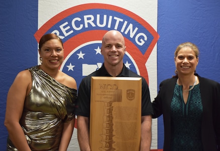 Three Soldiers smile and hold up an award plaque in front of an Army Recruiting Brigade backdrop