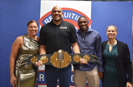 Two male Army Soldiers smile while holding a gold belt.  On either side are smiling female Soldier award presenters.