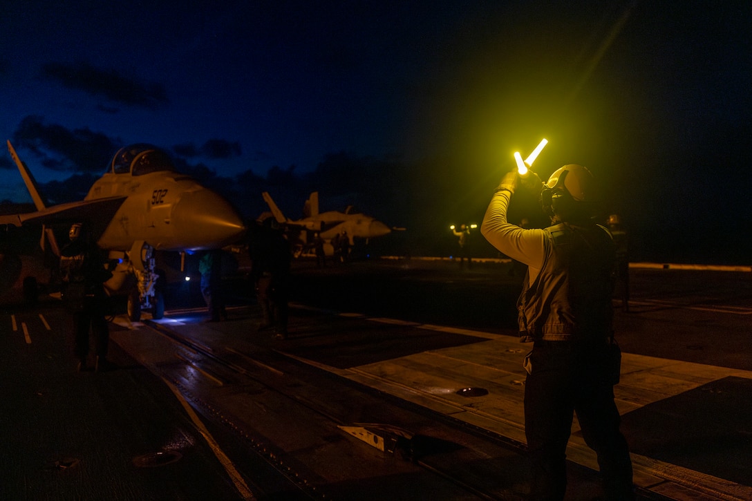 Sailors holding yellow marshalling wands direct a military aircraft on the flight deck of a Navy ship at night.