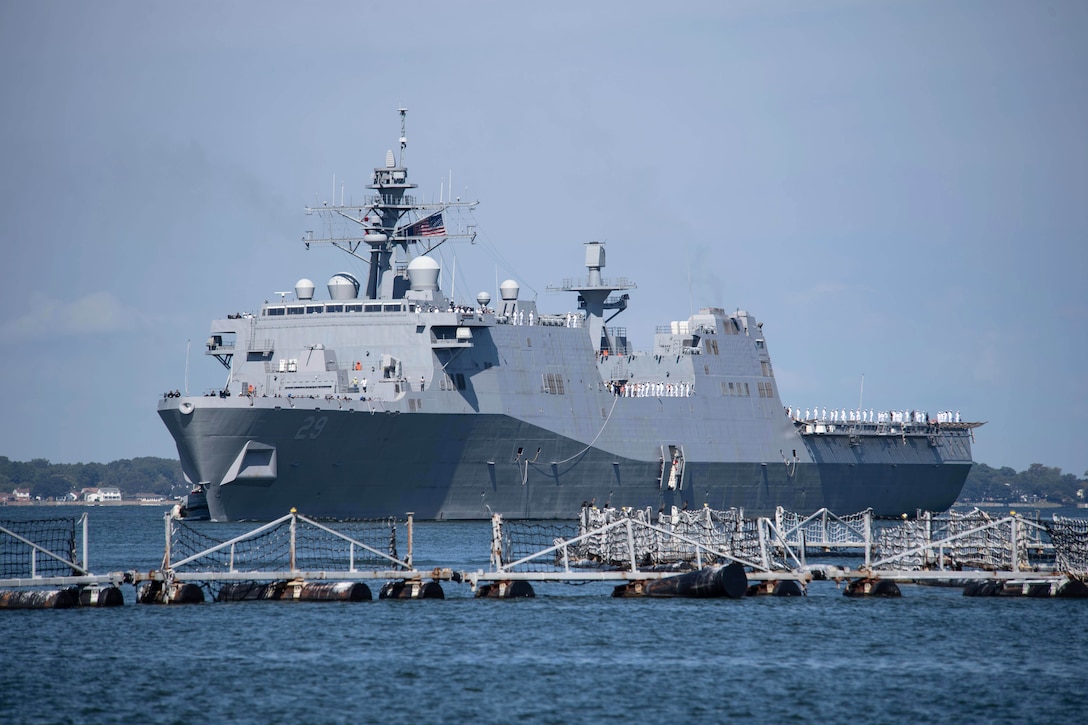 Dozens of sailors stand watch aboard a ship as it approaches a pier.