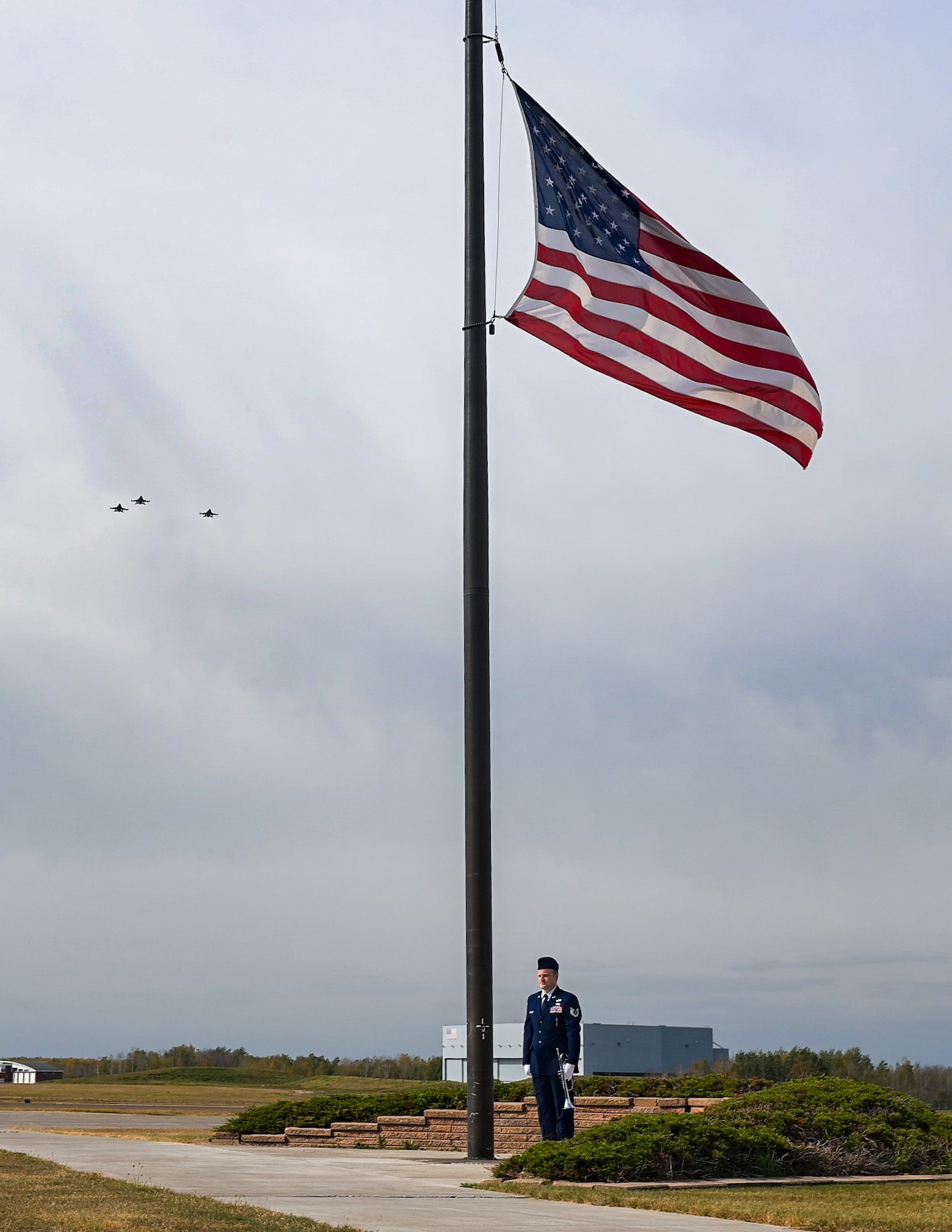 U.S. Air Force Tech. Sgt. Nathan Windus, a munitions systems specialist with the 148th Fighter Wing, Minnesota Air National Guard stands post after playing TAPS while three 179th Fighter Squadron F-16 Fighting Falcons, fly in the Missing Man Formation at a ceremony dedicating a memorial wall at the 148th Fighter Wing in Duluth, Minn. Oct. 5, 2024. The newly completed memorial wall honors 21 fallen service members who were lost while conducting their military duties over the wing’s 76-year history. (U.S. Air National Guard photo by Staff. Sgt. Tylin Rust)