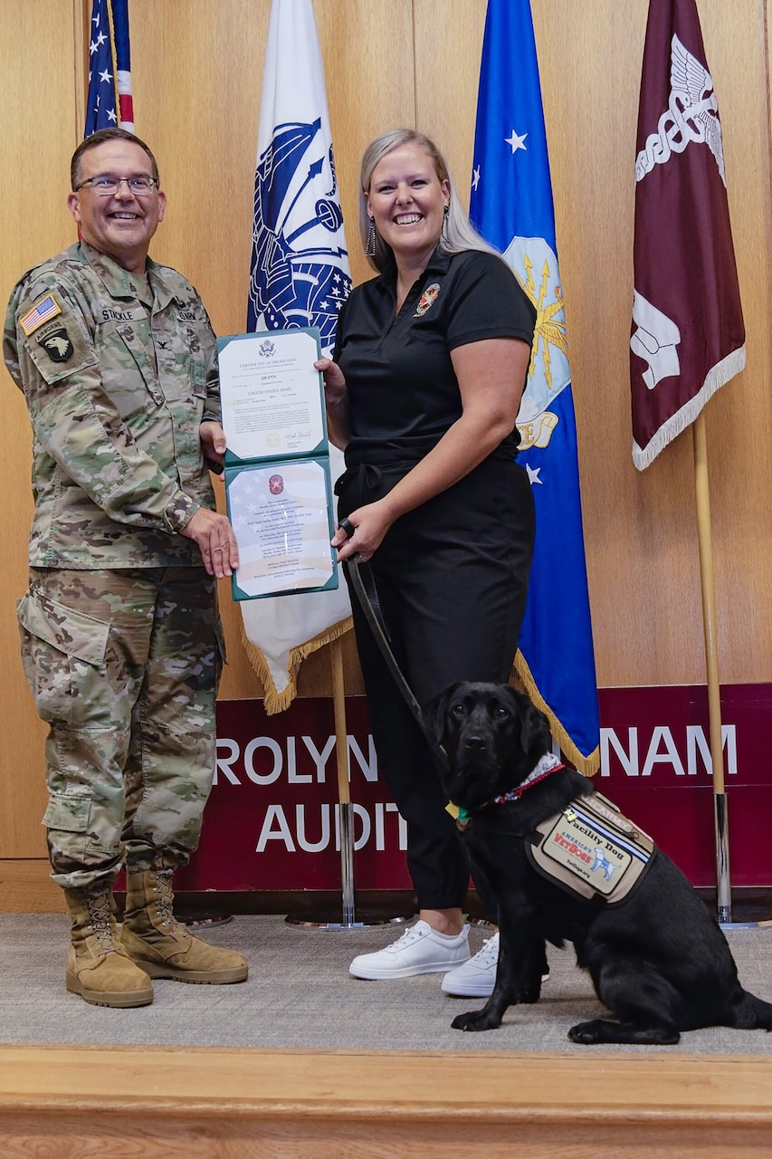A uniformed service member and a person in civilian clothing hold an award as a military dog sits next to them.