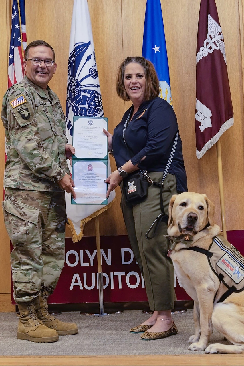 A uniformed service member and a person in civilian clothing hold an award as a military dog sits next to them.