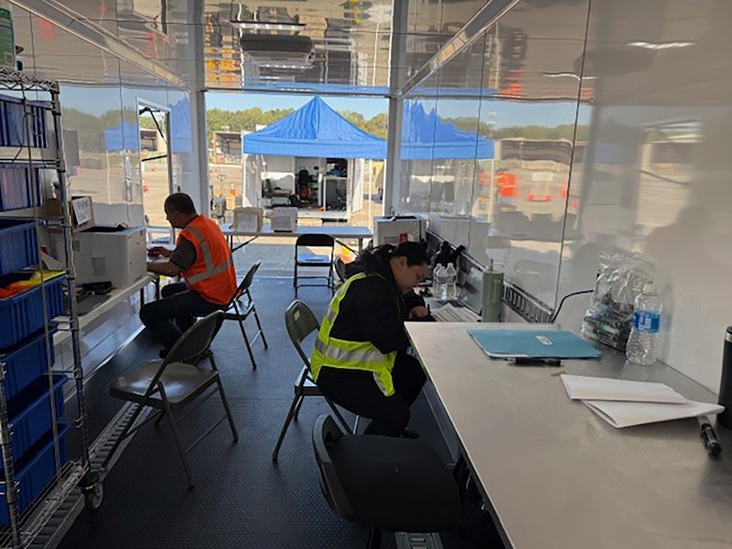 People in reflective vests work seated at desks inside a mobile trailer.