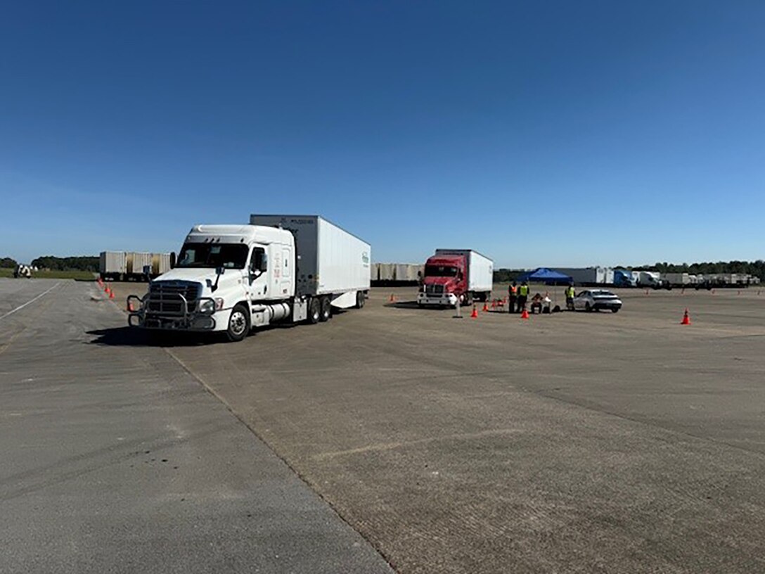 Tractor trailers are parked outside in a parking lot with people walking around in reflective vests.