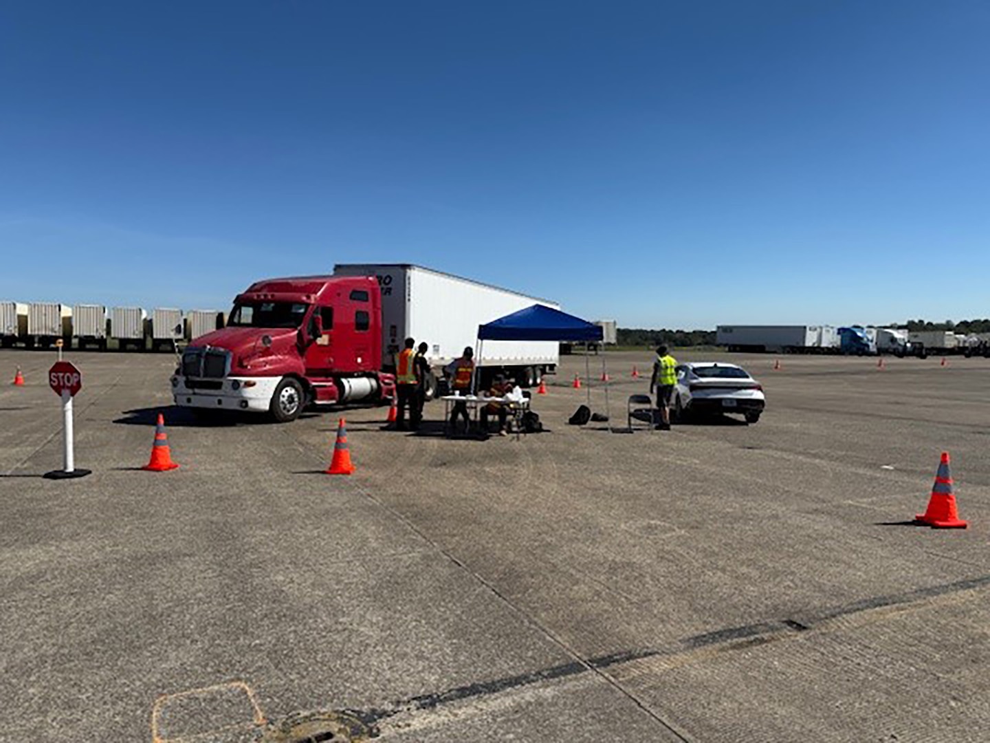 Tractor trailers are parked outside in a parking lot with people walking around in reflective vests.