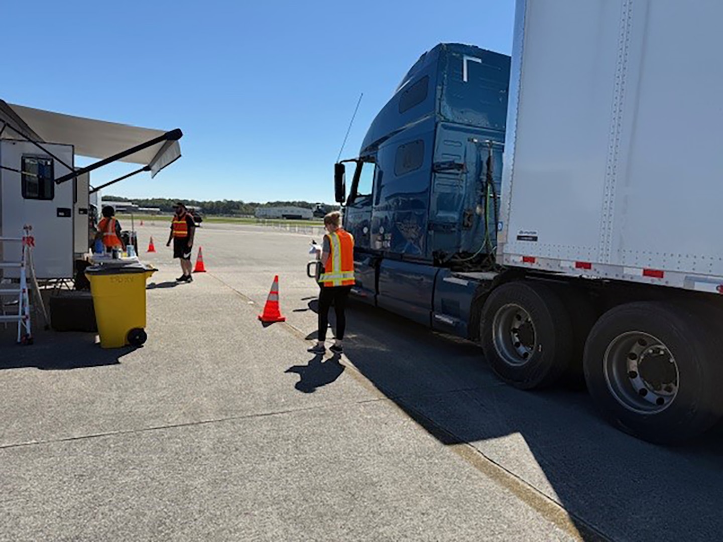 Tractor trailers are parked outside in a parking lot with people walking around in reflective vests.