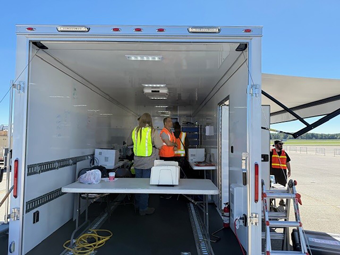 People in reflective vests stand inside an incident support base trailer that is parked outside in a parking lot.