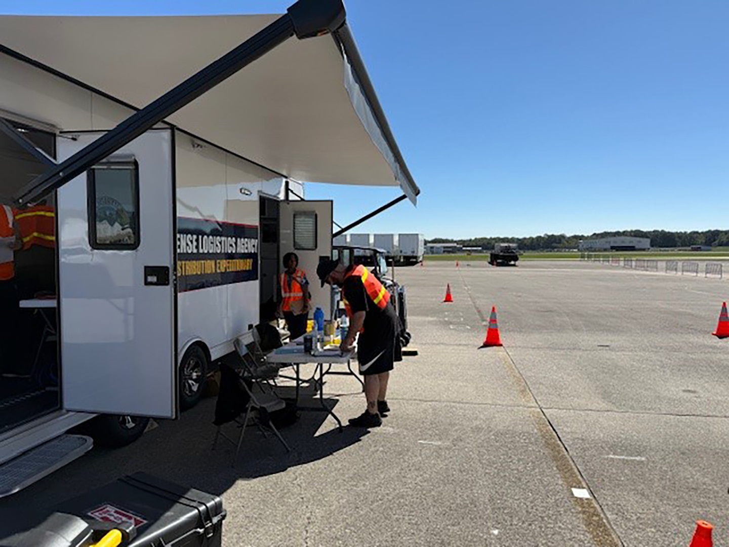 People in reflective vests write on papers on a table positioned outside of an incident support base trailer that is parked outside in a parking lot.