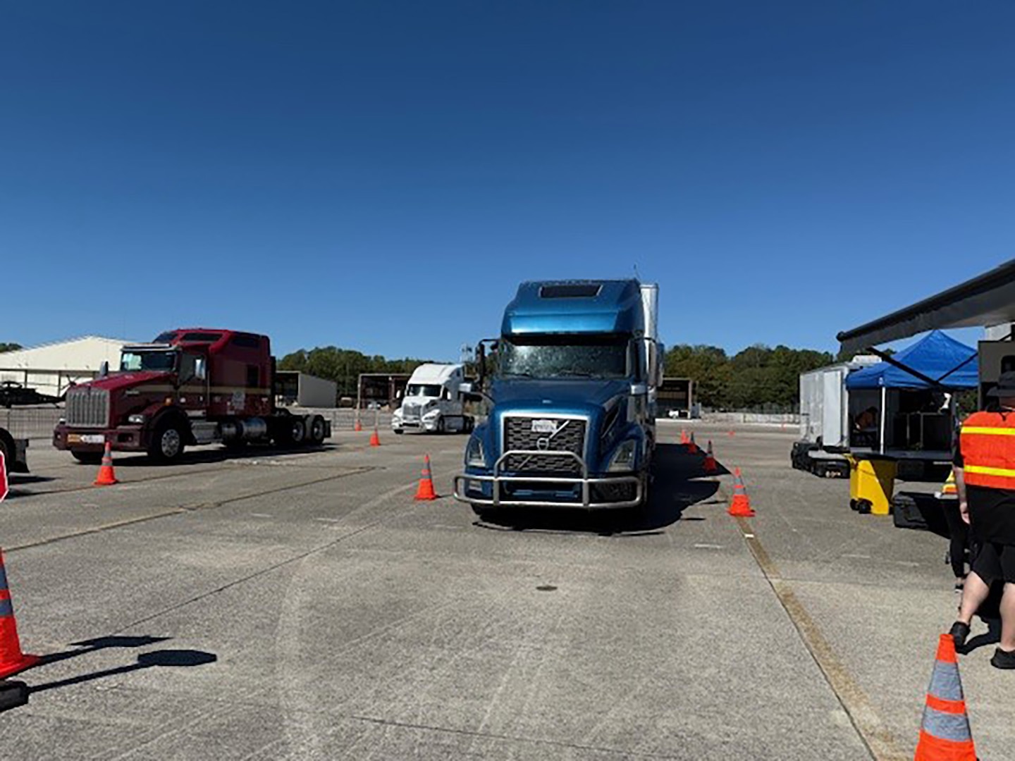 Tractor trailers are parked outside in a parking lot with people walking around in reflective vests.