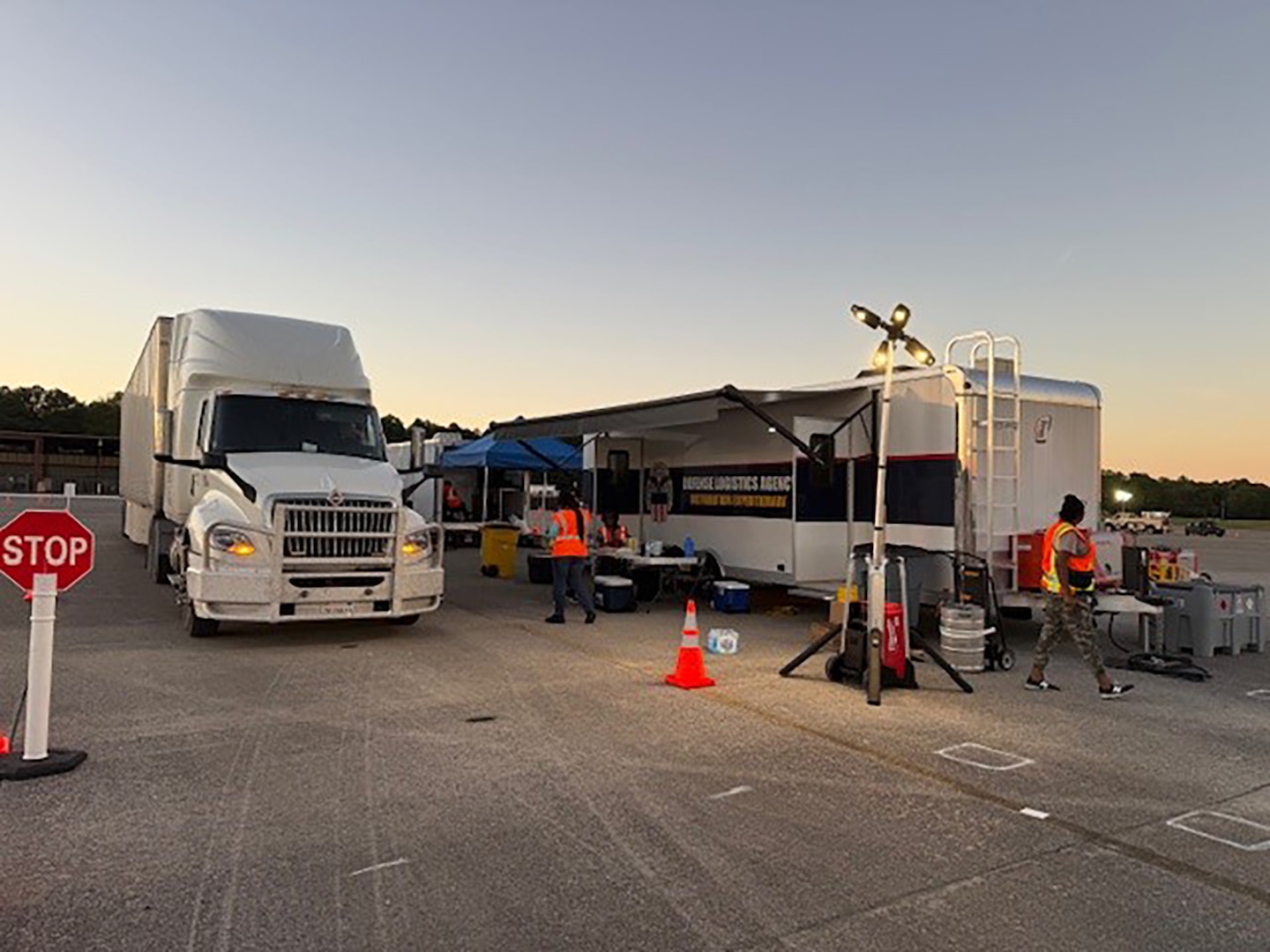 A tractor trailer and a incident support base shelter are outside in a parking lot with people walking around in reflective vests.