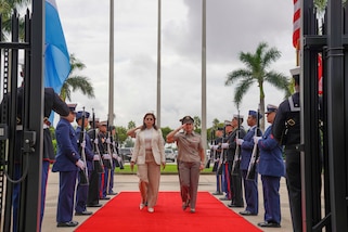 A female military officer and a woman in a white suit walk a red carpet flanked by military personnel standing in formation with ceremonial rifles.