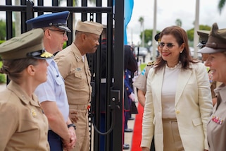 Military personnel greet a woman in a white suit.