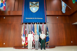 A woman in a white suit and two military personnel stand in front of flags for a group photo.