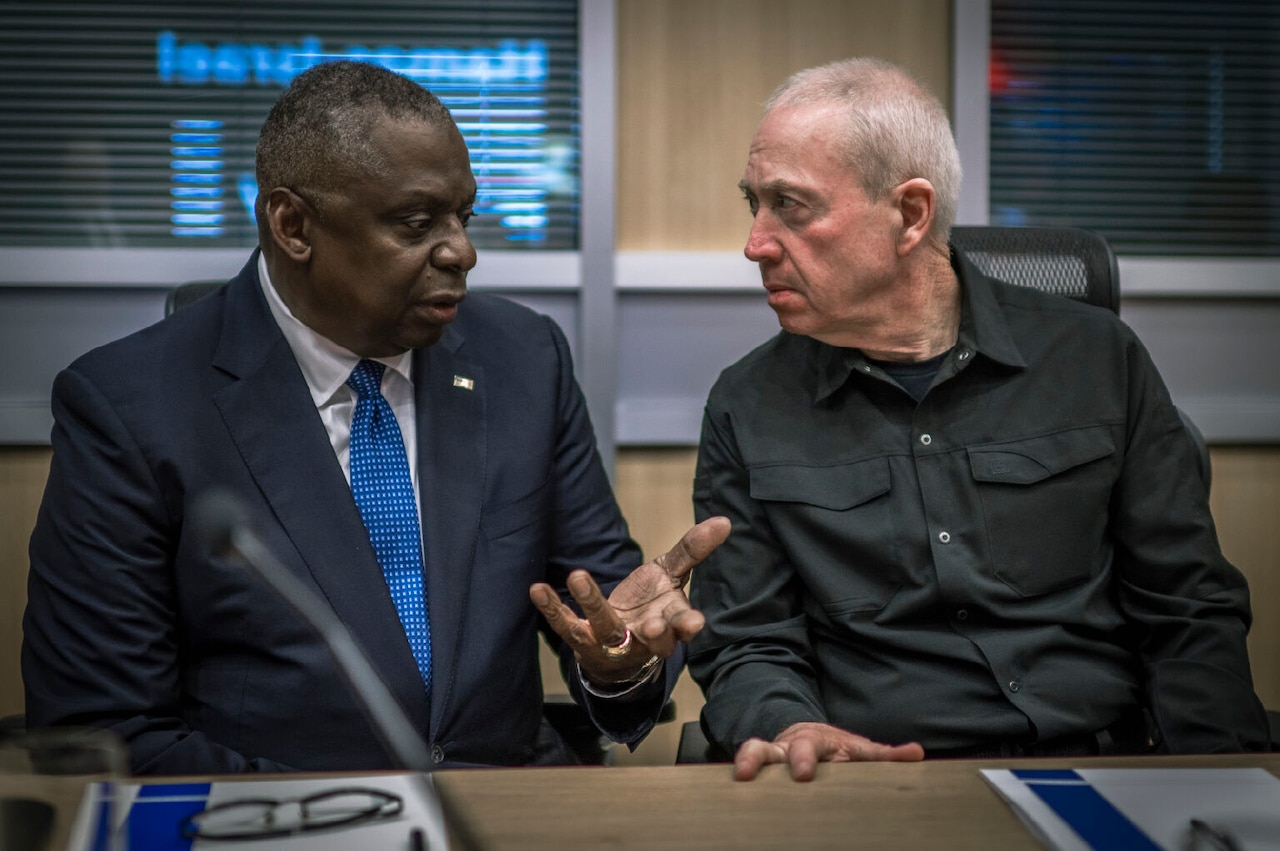 Two men in civilian attire talk while seated at a table.