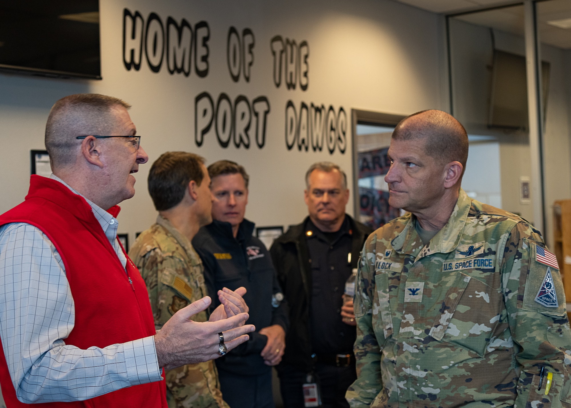 A man in uniform speaks to a man in a room with three men in the background and large text on the wall that reads HOME OF THE PORT DAWGS.