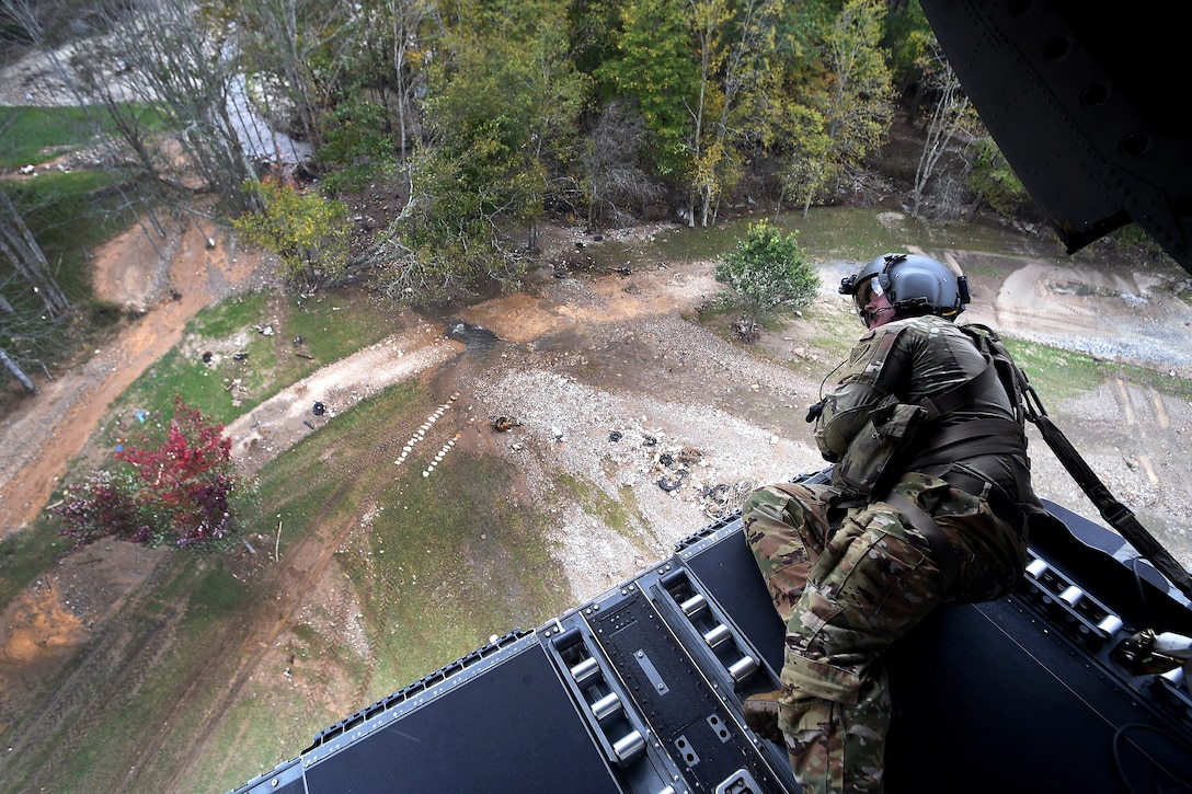 A soldier looks over a debris-strewn field from the back of a low-flying open helicopter.